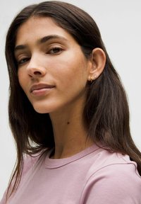 Camiseta de algodón rosa con escote redondo, llevada por una persona con cabello largo y oscuro. Pendientes de aro dorados pequeños visibles. Fondo neutro.