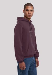 Bordeauxfarbener Hoodie aus weichem Stoff mit Kängurutasche, Kapuze mit Kordelzug und dezentem Logodetail auf der Brust, kombiniert mit blauen Jeans.