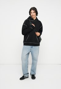 Sort hættetrøje med hvide syninger, parret med løse lyseblå jeans og sorte sneakers. Modellen poserer med en taske.
