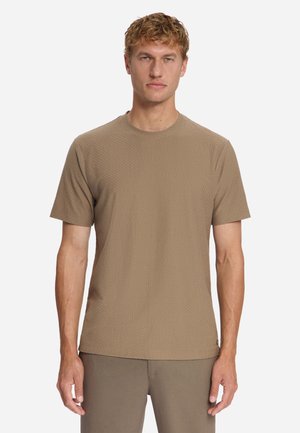 Man met kort krullend haar, gekleed in een gestructureerd beige shirt met korte mouwen en bijpassende beige broek, staand tegen een effen achtergrond.