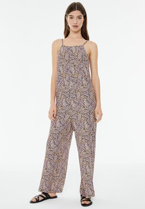 Jumpsuit s cvjetnim uzorkom, s podesivim trakama, lepršavim hlačama širokih nogavica i laganim materijalom u ljubičastim, ružičastim i žutim tonovima.