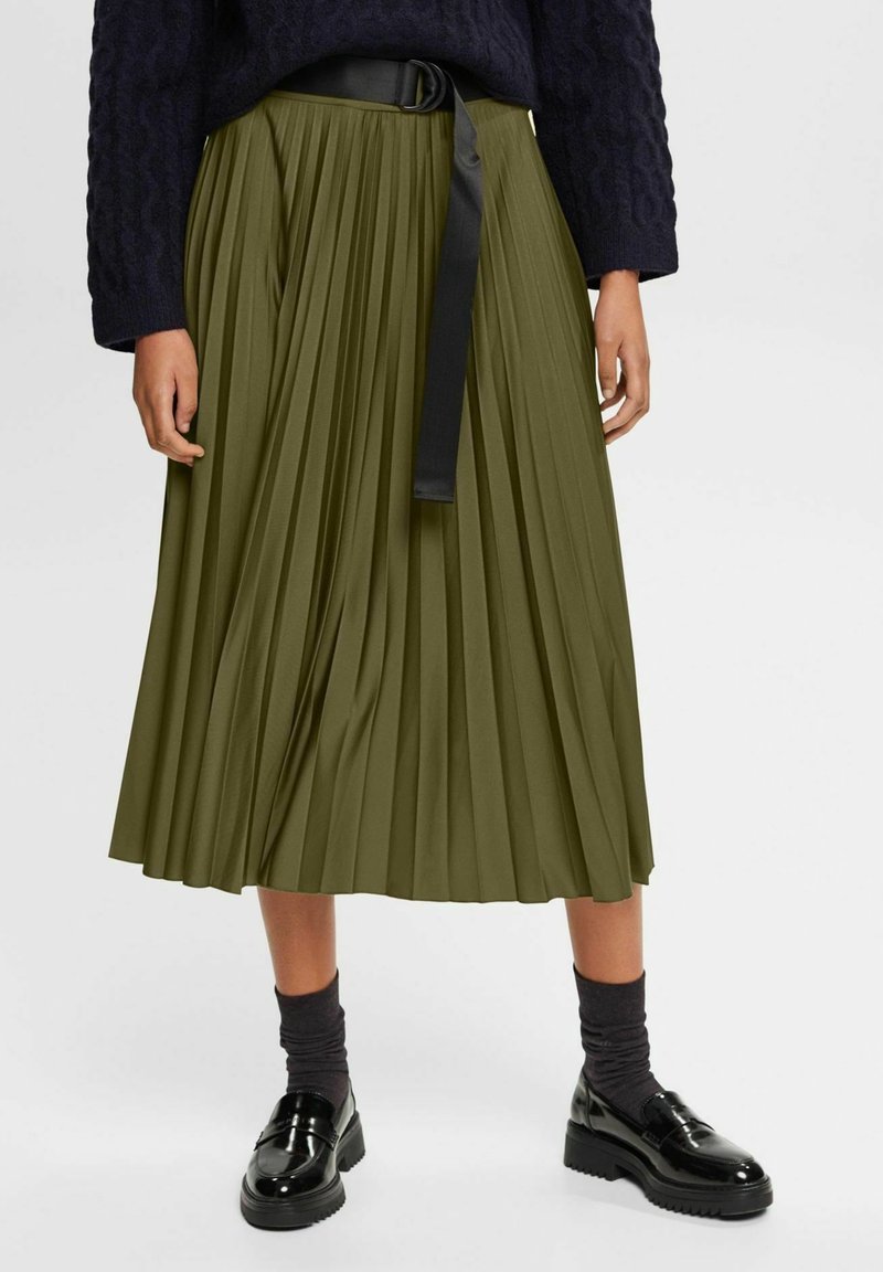 Femme portant une jupe midi plissée vert olive avec une ceinture noire, des chaussettes foncées, des mocassins noirs brillants et un pull texturé bleu marine.