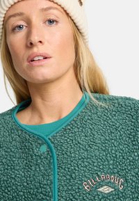 Vihreä fleece-takki, jossa on teksturoitu pinta, ribattu teal-värinen pyöreä kaulus ja vaaleanpunainen Billabong-logo brodeerattuna rintaan.