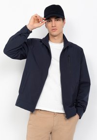 Veste bleu marine fabriquée dans un matériau lisse, dotée d'une poche poitrine zippée, de poches latérales et d'un col montant. Le modèle porte une casquette noire.