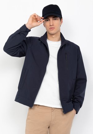 Giacca blu navy realizzata in materiale liscio, dotata di una tasca sul petto con zip, tasche laterali e colletto alto. Il modello indossa un cappellino nero.