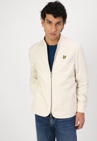Lys beige jakke med lynlås og stående krave. Har et lille gult logo. Bæres over en mørkeblå skjorte og kombineres med blå jeans.