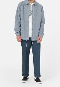 Pruhovaná košile s knoflíky v modré a bílé barvě, kombinovaná s tmavými modrými kalhotami s tkaničkou. Černé slip-on boty dotvářejí tento outfit.