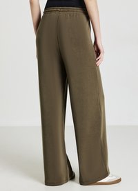 Weite Hosen aus olivgrünem Stoff mit glatter Textur. Mit einem elastischen Bund und seitlichen Schlitzen, kombiniert mit hellfarbigen Schuhen.