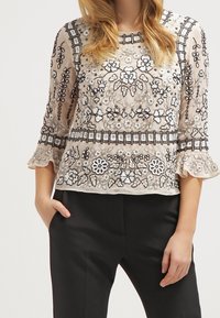 Femme portant un haut beige transparent avec une broderie florale noire et blanche, et un pantalon noir, debout avec une main dans la poche.