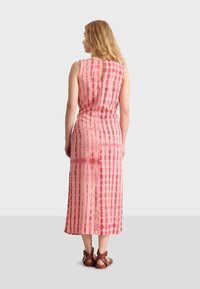 Un largo vestido rosa con un patrón de tie-dye, que presenta un diseño sin mangas, un escote en forma de ojo de cerradura y una silueta fluida. Combinado con sandalias marrones.