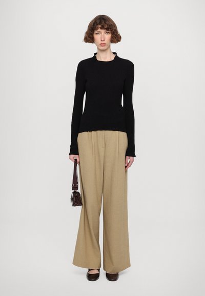 Black long-sleeve knit top with a subtle textured finish, paired with loose beige trousers. A small brown handbag is held in the right hand.