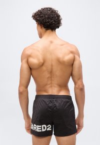 Hombre musculoso con cabello rizado y oscuro de espaldas, lleva pantalones cortos negros con el texto "DSQUARED2" en blanco en la pierna izquierda.