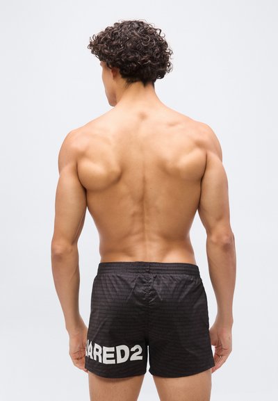 Homme musclé aux cheveux bouclés et foncés, vu de dos, portant un short noir avec l'inscription "DSQUARED2" en texte blanc sur la jambe gauche.