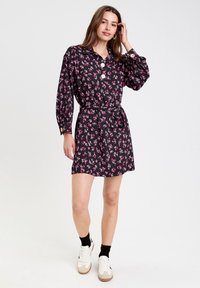 Vestido negro con estampado floral que cuenta con mangas largas, cuello abotonado y cintura ceñida con un cinturón. La tela tiene una textura suave con patrones florales en tonos rosas.