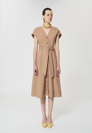 Femme portant une robe beige longueur genou avec des boutons et une ceinture nouée à la taille, accessoirisée de boucles d'oreilles dorées, un collier doré épais et des escarpins jaunes à motifs.