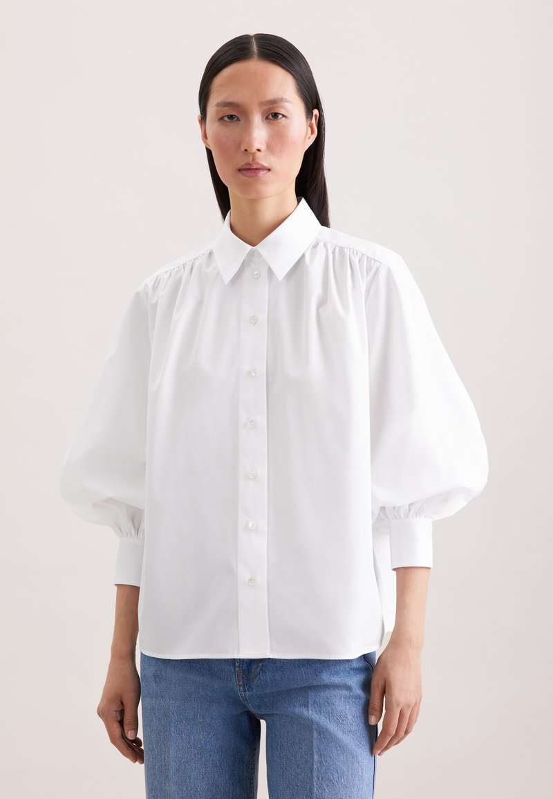 Femme portant un chemisier blanc boutonné avec des manches bouffantes et un jean bleu, debout devant un fond clair uni.