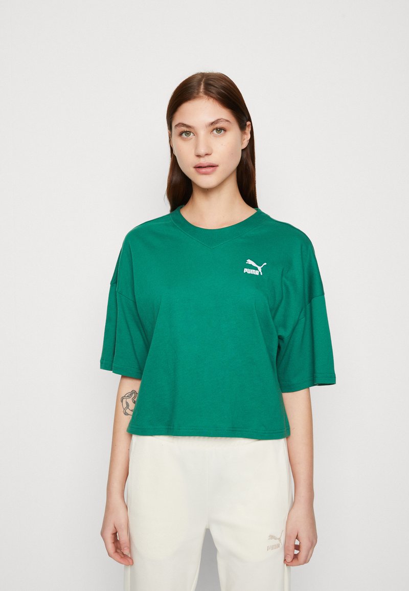 T-shirt verde in cotone con maniche corte e larghe, colletto rotondo, e logo bianco Puma sul lato sinistro. Indossato con pantaloni chiari.