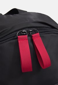 Sac à dos noir avec une texture lisse, une fermeture éclair avec des tirettes en tissu rouge, et un design arrondi. Agrémenté de détails brodés.