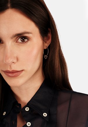 Pendientes de aro metálicos con diseño en forma de corazón, con un acabado pulido en plata. El modelo lleva una camisa oscura abotonada.