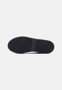 Σόλα ενός μαύρου παπουτσιού Lacoste που δείχνει το μοτίβο πέδιλου ψαροκόκκαλου και το λογότυπο της Lacoste σε λευκό φόντο.