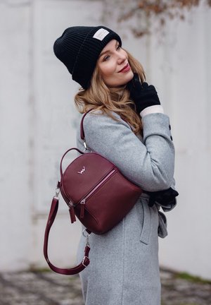 Burgundská kožená batoh s zaobleným tvarem, dvěma zipy a nastavitelného popruhem. Model nosí šedý kabát a černou pletenou čepici.