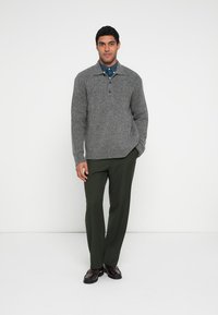 Pelēks adīts džemperis ar augstu apkakli un divu pogu aizdari, kombinēts ar tumši zaļām biksēm un brūnām kurpēm. Gluda tekstūra un piegulošs dizains.