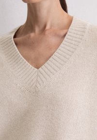 Beige v-hals gebreid trui met geribbelde nek, met een gestructureerd oppervlak en een ontspannen pasvorm. Geen zichtbare patronen of hardwaredetails.