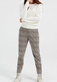 Kvinna i en vit stickad tröja, rutiga tapered byxor och off-white snörade ankelboots, stående med armarna i kors.