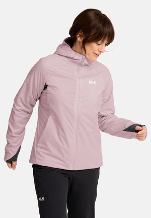 Vrouw draagt een lichtpaarse capuchonjas en zwarte broek met zichtbare Jack Wolfskin-logo's, kijkt naar beneden en glimlacht licht.