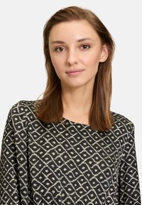 Schwarze Bluse mit einem abstrakten weißen Muster. Verfügt über einen runden Ausschnitt und lange Ärmel, hergestellt aus leichtem Stoff. Glatte Textur.
