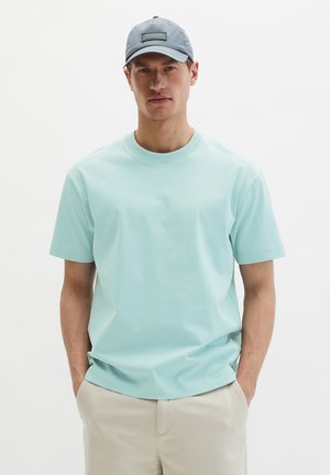 Homme portant un t-shirt bleu clair à col rond, un pantalon beige et une casquette de baseball grise, les mains dans les poches, devant un fond uni.