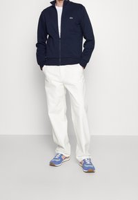 Giacca zip con colletto alto di colore blu navy, t-shirt bianca sotto, pantaloni bianchi e scarpe sportive blu con dettagli arancioni.