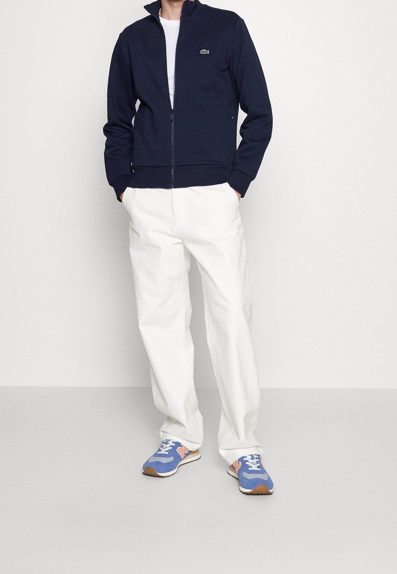 Giacca zip con colletto alto di colore blu navy, t-shirt bianca sotto, pantaloni bianchi e scarpe sportive blu con dettagli arancioni.