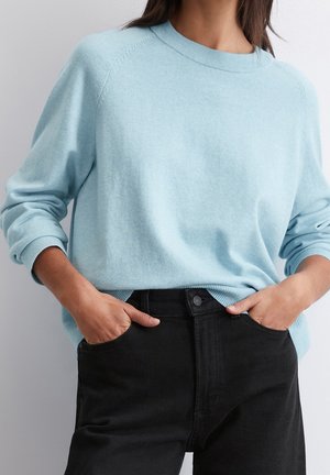 Vrouw draagt een lichtblauwe trui met lange mouwen, gestopt in een zwarte high-waisted jeans, met handen in de voorzakken.
