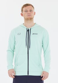 Lys mintgrøn hættetrøje med lynlås, lavet af blødt stof, har en frontlynlås, to lommer og kontrasterende marineblå snore og detaljer.