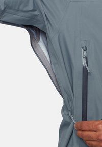 Hånd trækker lynlåsen på blå jakke under arm ventilation åbning, der viser lys grå netforing indeni.