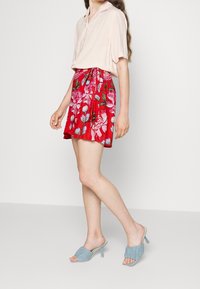 Blusa rosa chiaro con maniche corte abbinata a una gonna rossa con stampa floreale. La modella indossa sandali con tacco azzurri.