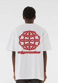 Valge puuvillane T-särk, mille seljal on suur punane graafika abstraktsete kujunditega ja tekst "mjgonzales" paksus punases fondis.