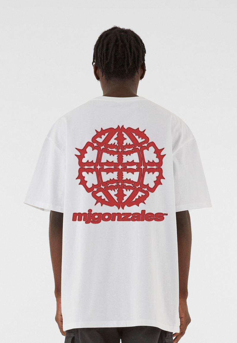 Valge puuvillane T-särk, mille seljal on suur punane graafika abstraktsete kujunditega ja tekst "mjgonzales" paksus punases fondis.