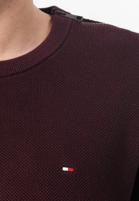 Pull en tricot bordeaux avec un motif texturé, doté d'une fermeture éclair noire à l'épaule et d'un petit logo brodé en rouge, blanc et bleu.