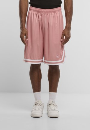 Homme portant un short de basket rose avec une bordure blanche, des chaussettes blanches et des baskets blanches, debout devant un fond uni.