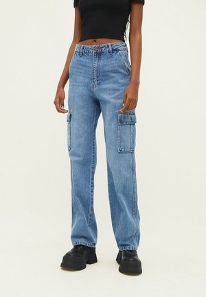 Kvinde iført lyseblå denim cargo jeans med store sidelommer, en sort crop-top og sorte platformsko, stående mod en ensfarvet baggrund.