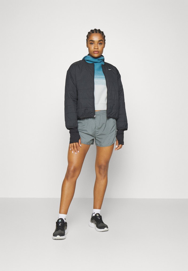 Schwarze gesteppte Bomberjacke über einem blau-grauen gemusterten Pullover, kombiniert mit grauen Shorts und schwarzen Sportschuhen mit weißen Akzenten.