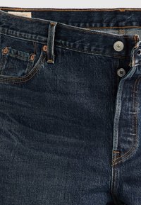 Jean en denim bleu foncé avec poches avant, passants de ceinture, bouton métal visible et surpiqûres orange détaillées.