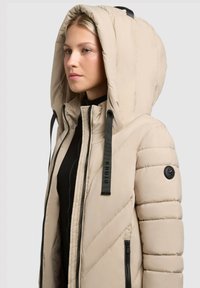 Beige pufjakke med en stor hætte, ziplukning og sorte detaljer. Tekstureret materiale med diagonale syninger og logo-accenter.