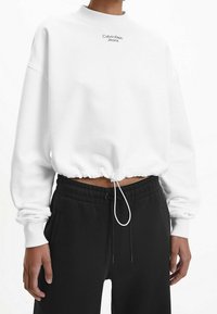 Sudadera blanca corta con cuello acanalado, dobladillo elástico y cordón ajustable. Presenta un pequeño logotipo negro "Calvin Klein Jeans" en el frente.