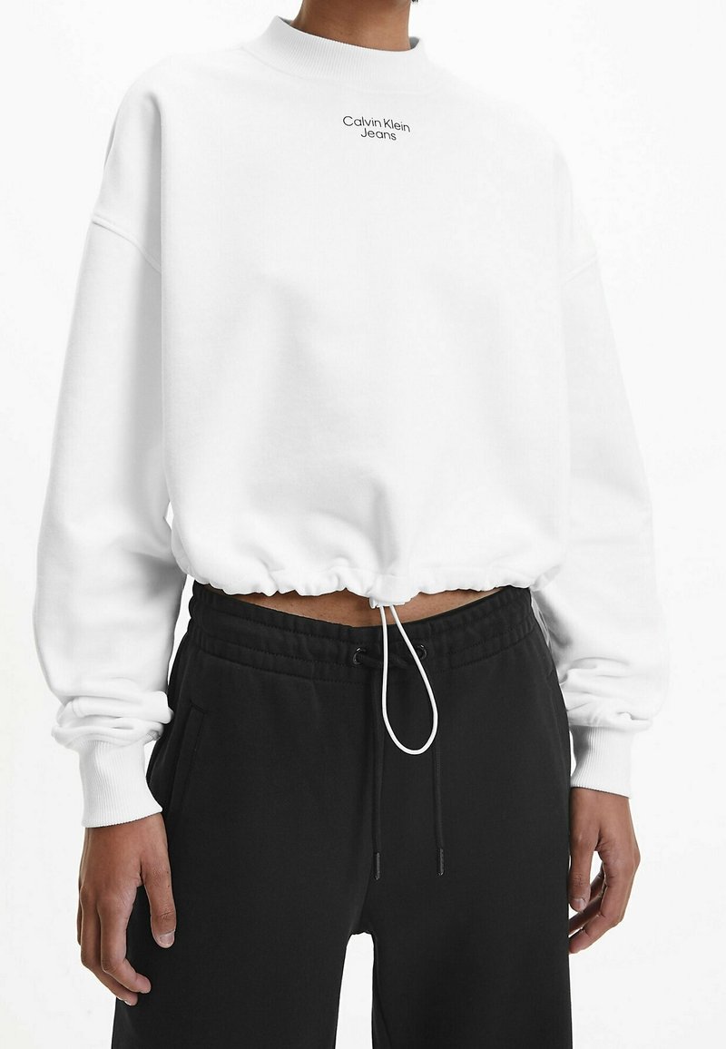 Sudadera blanca corta con cuello acanalado, dobladillo elástico y cordón ajustable. Presenta un pequeño logotipo negro "Calvin Klein Jeans" en el frente.