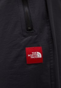 Mørkegrå stof med en subtil tekstur prydes af en sort lynlås og en rød vævet etiket, der viser "THE NORTH FACE" i hvid.