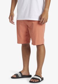 Shorts en corail en coton, avec des jambes droites et des poches latérales, assortis à des sandales noires à glissière avec un design rayé.