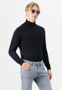 Schwarzer Rollkragenpullover mit eng anliegendem Design, kombiniert mit hellgrauen Jeans. Das Model trägt Sonnenbrillen; der Stoff wirkt glatt und figurbetont.